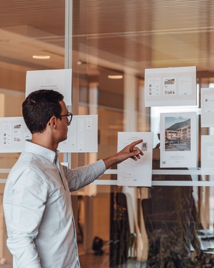 HMM – The Tourism Group Mann zeigt auf Designentwürfe, die an Glaswand in Büro geklebt sind